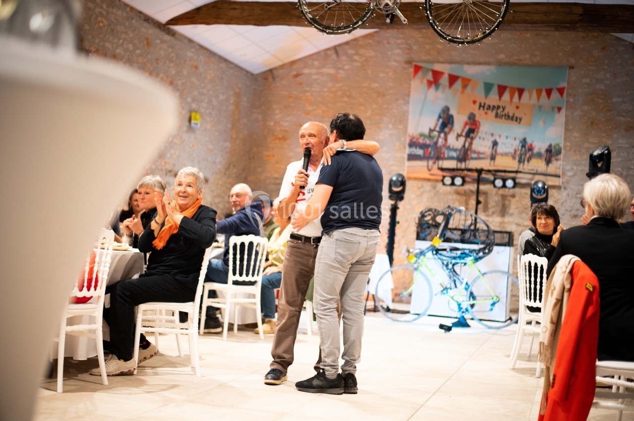 Deux hommes s'enlacent lors d'une fête d'anniversaire dans une salle décorée, avec des invités assis applaudissant.