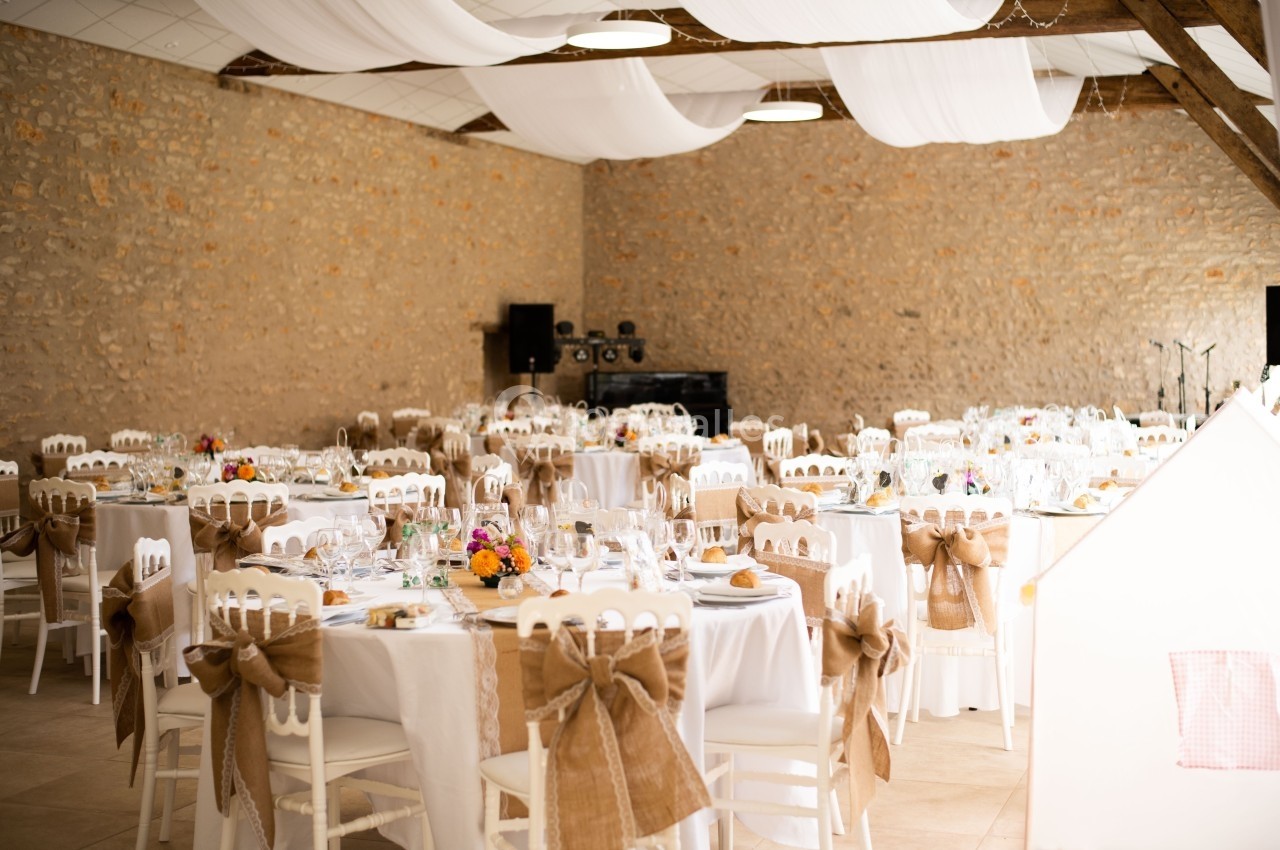Salle de réception décorée avec des tables rondes, nappes blanches, chaises ornées de nœuds en toile de jute et fleurs…