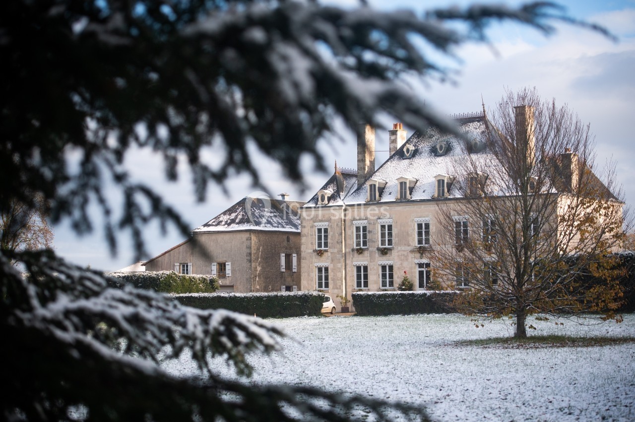Manoir en pierre entouré d'un jardin enneigé, partiellement encadré par des branches d'arbres au premier plan.