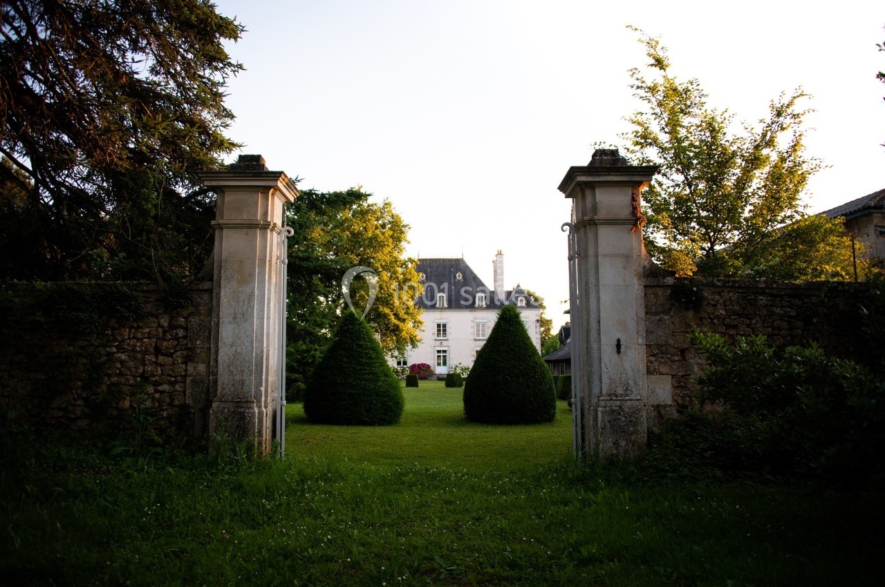 Entrée d'un jardin avec des piliers en pierre, menant à un bâtiment ancien entouré de verdure.