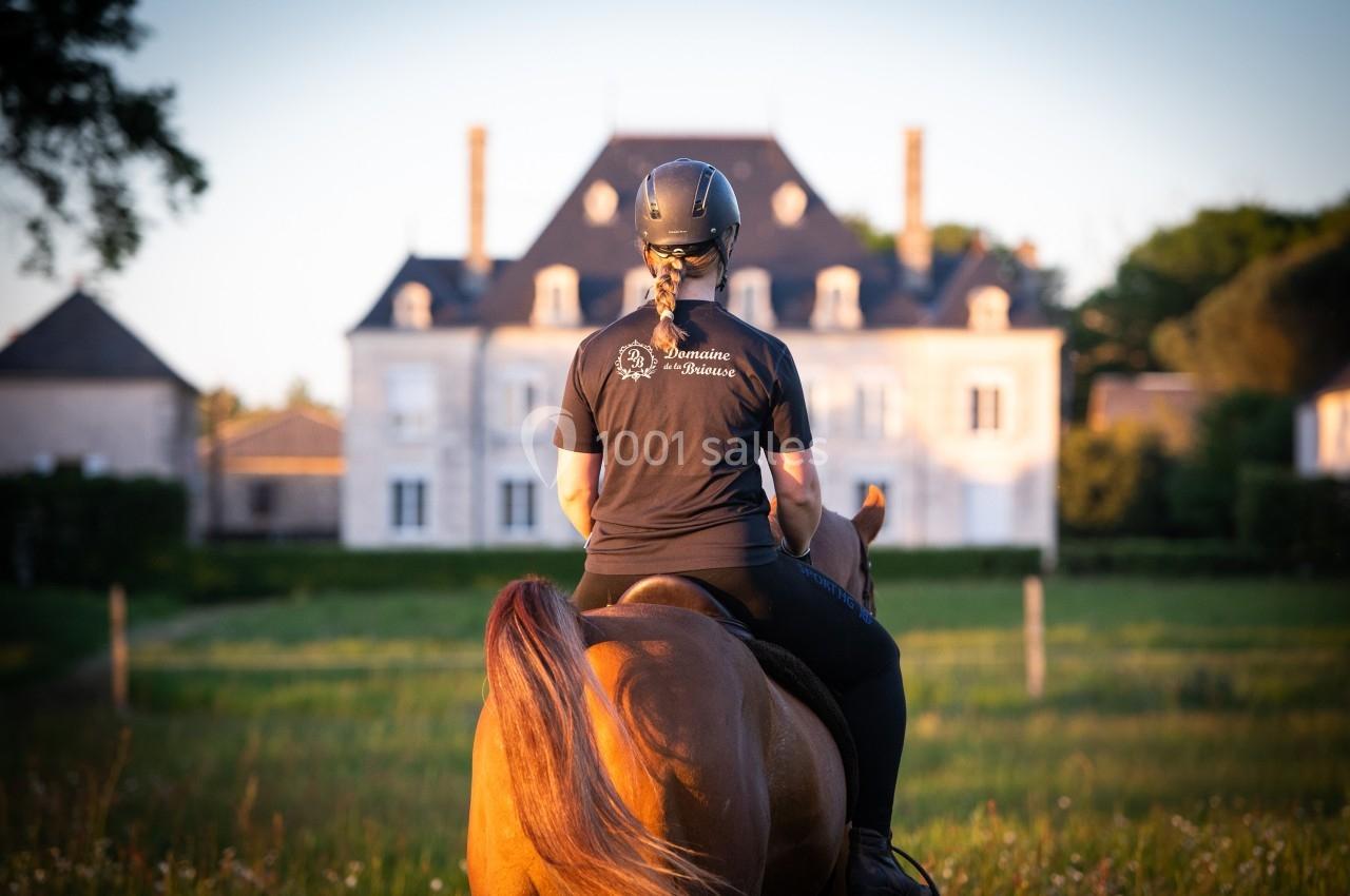 ACTIVITES AU DOMAINE Une cavalière vue de dos, montant un cheval dans un pré, avec un manoir en arrière-plan au coucher du soleil.