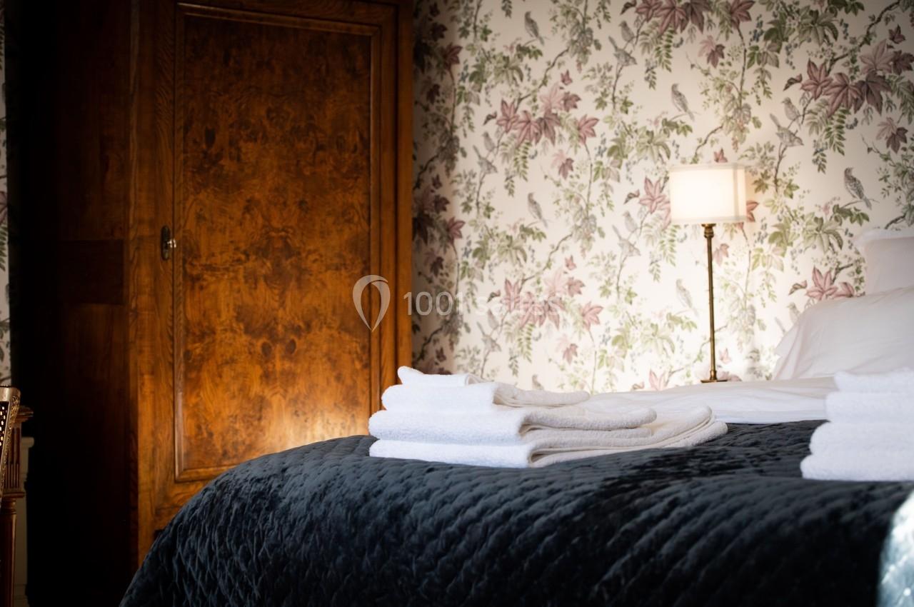 CHAMBRE D'HOTES Lit avec couvre-lit sombre, serviettes blanches pliées, armoire en bois et mur tapissé de motifs floraux en arrière-plan.