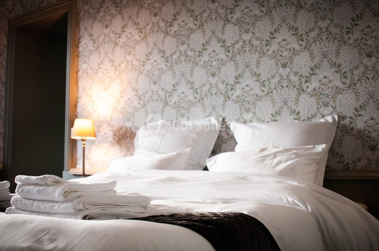 CHAMBRE D'HOTES Lit double avec draps blancs, oreillers moelleux, serviettes pliées au premier plan et lampe allumée en arrière-plan.