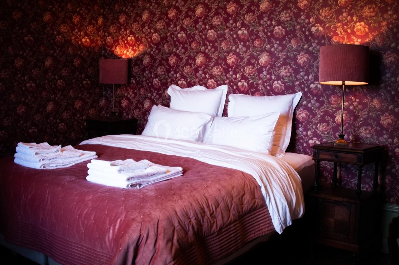 CHAMBRE D'HOTES Lit double avec draps blancs, couverture rouge et serviettes pliées, entouré de lampes et d'un papier peint floral.