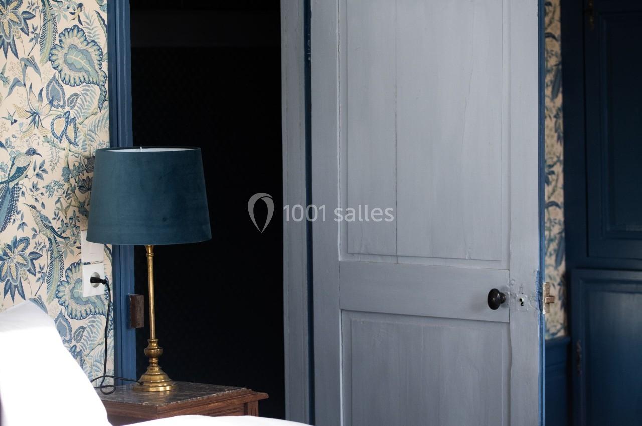 CHAMBRE D'HOTES Chambre avec lit, lampe sur table de chevet, mur tapissé à motifs floraux et porte entrouverte.