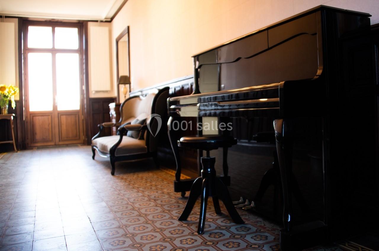 DOMAINE DE LA BRIOUSE Piano droit noir dans un salon lumineux avec carrelage ancien, fauteuils en bois et une fenêtre en arrière-plan.
