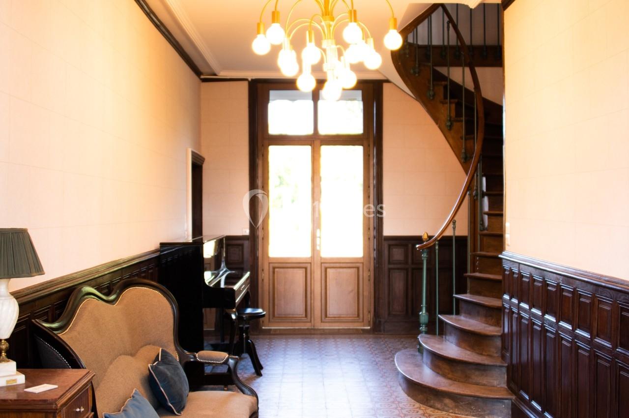 DOMAINE DE LA BRIOUSE Couloir lumineux avec carrelage ancien, canapé, piano, escalier en colimaçon et porte-fenêtre en arrière-plan.