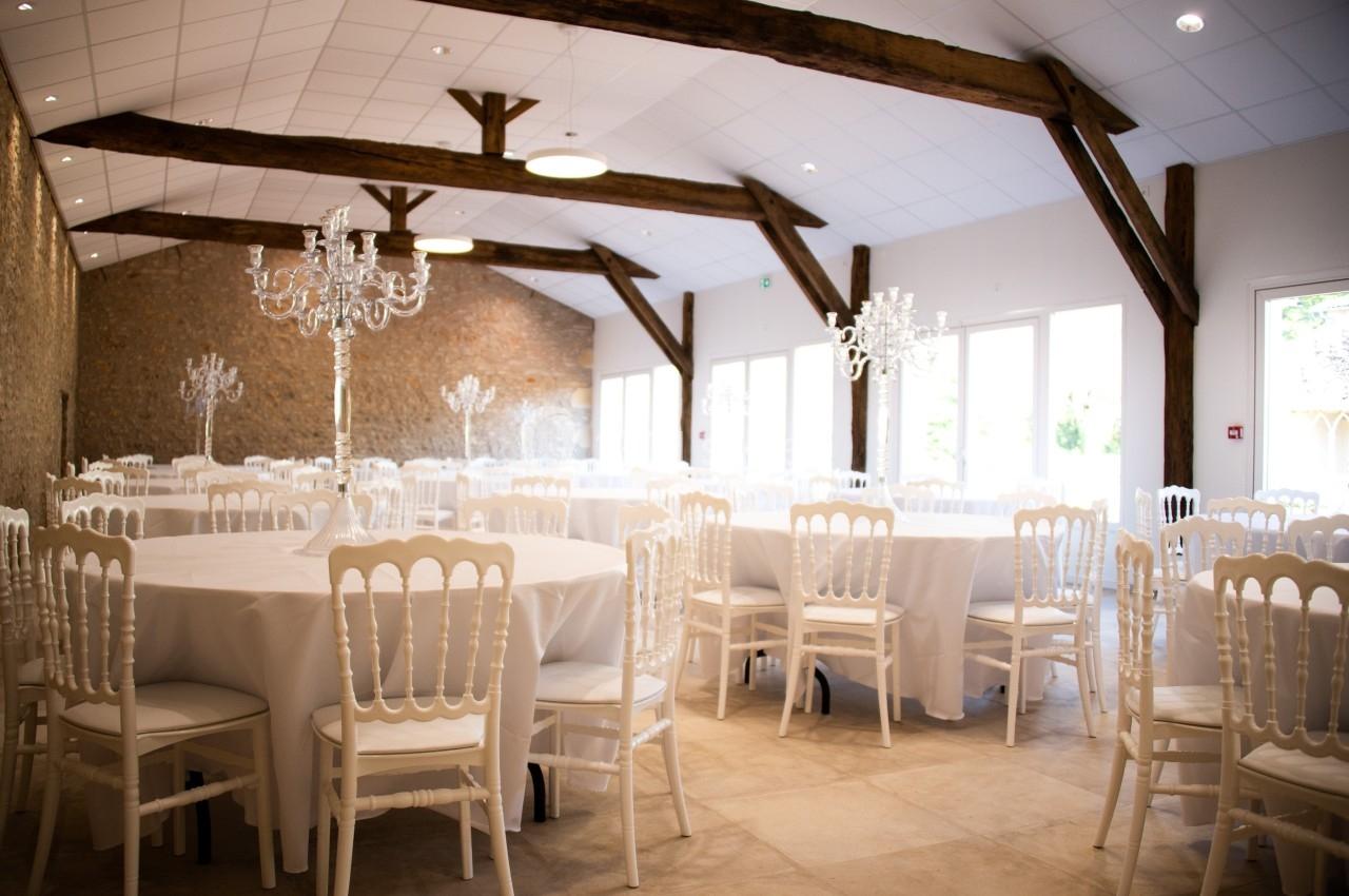 SALLE RIGOLETTO Salle de réception lumineuse avec des tables rondes dressées, des chaises blanches et des chandeliers élégants.