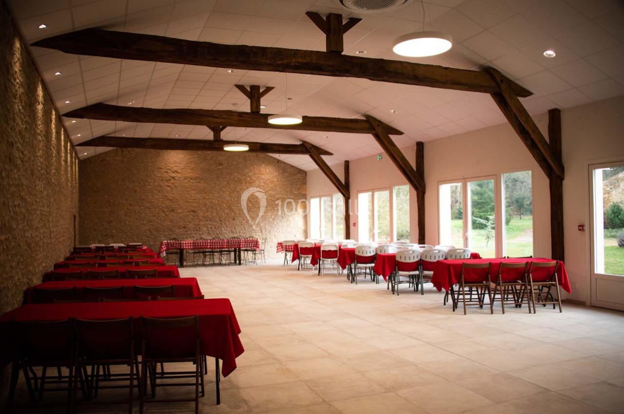 SALLE RIGOLETTO Salle spacieuse avec murs en pierre, poutres apparentes, tables rouges et grandes baies vitrées donnant sur un jardin.