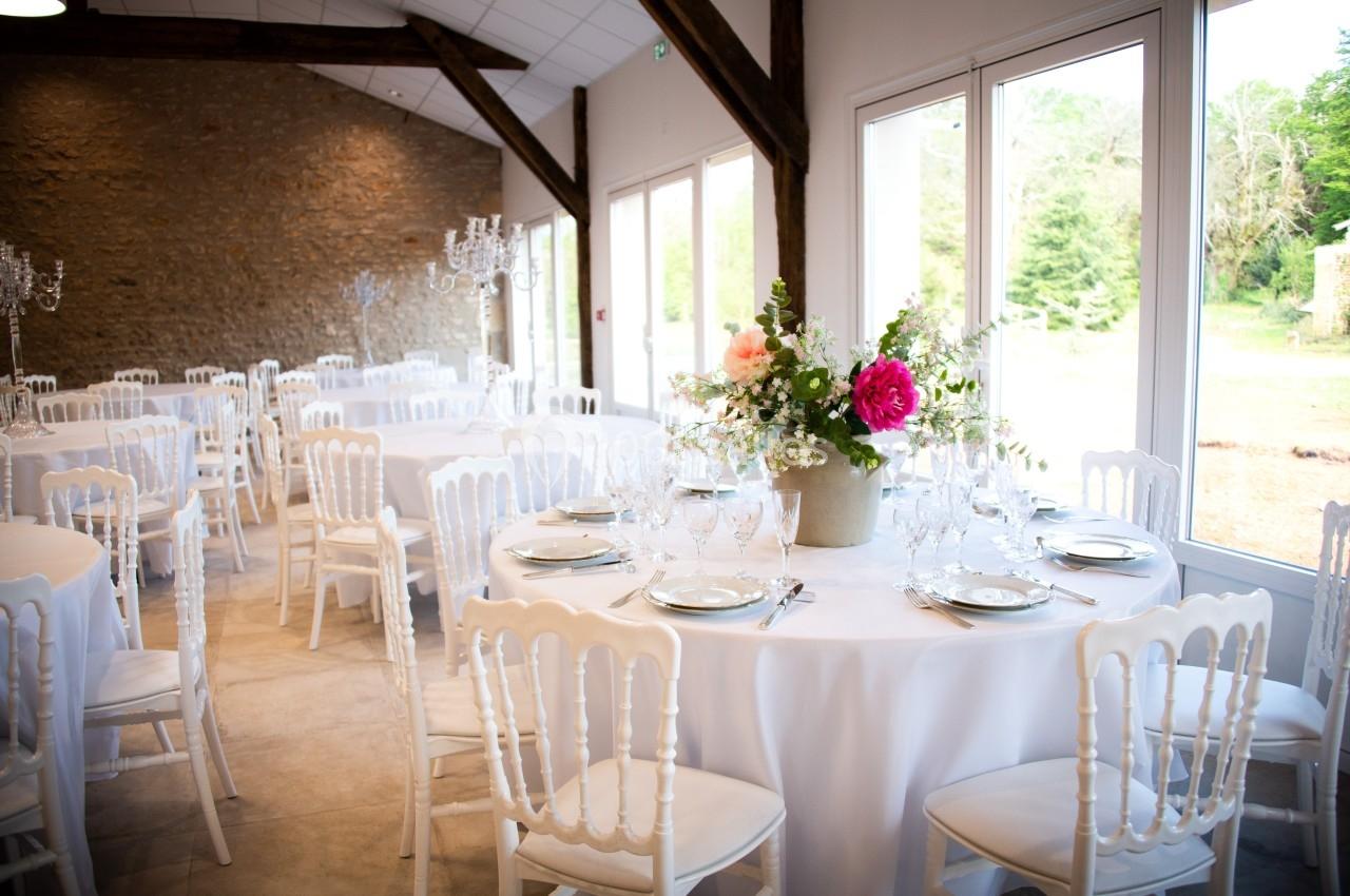 SALLE RIGOLETTO Salle de réception lumineuse avec tables rondes dressées, chaises blanches et centre de table floral.