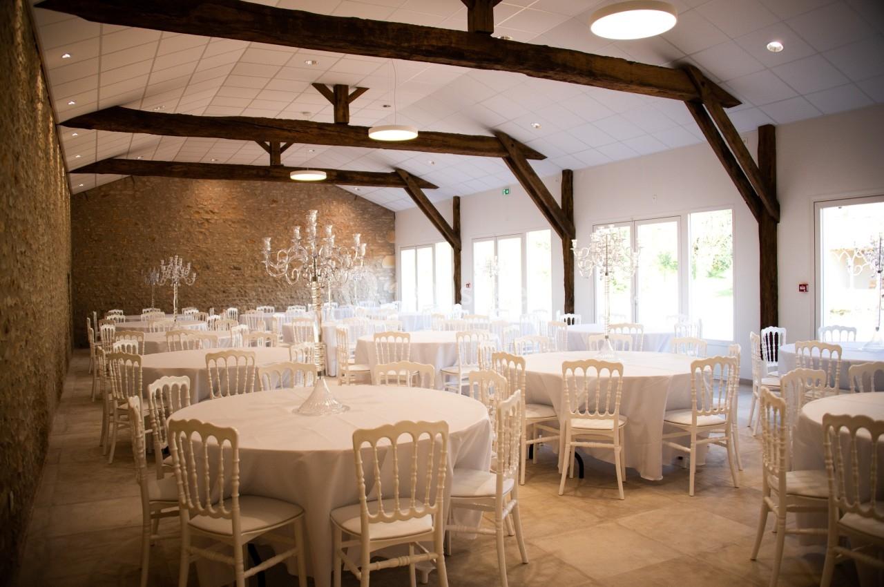 Salle de réception lumineuse avec tables rondes dressées de nappes blanches et chaises assorties, poutres apparentes.
