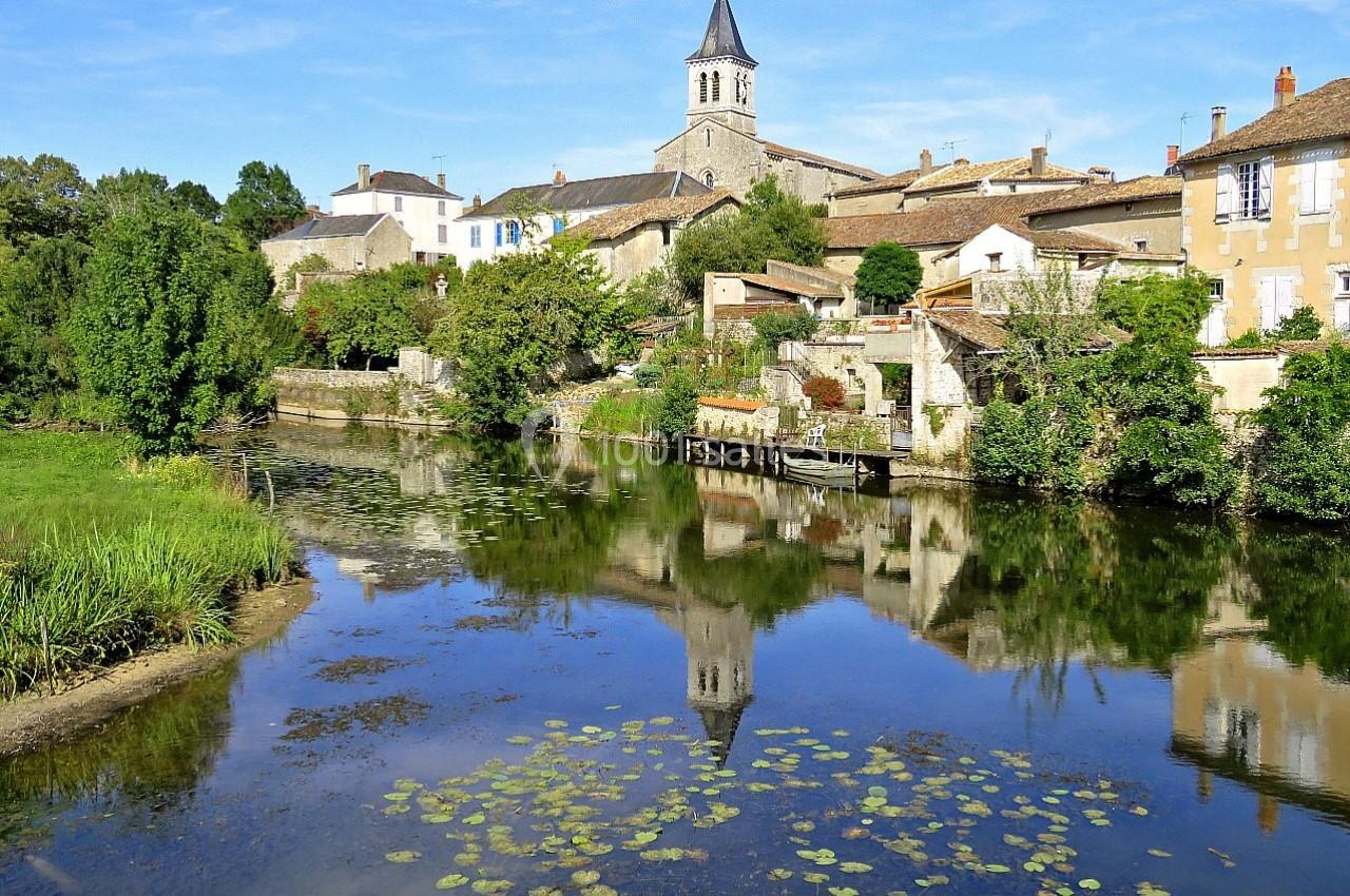 SANXAY Vue d'un village avec une église, des maisons en pierre et une rivière bordée de végétation, reflet dans l'eau calme.