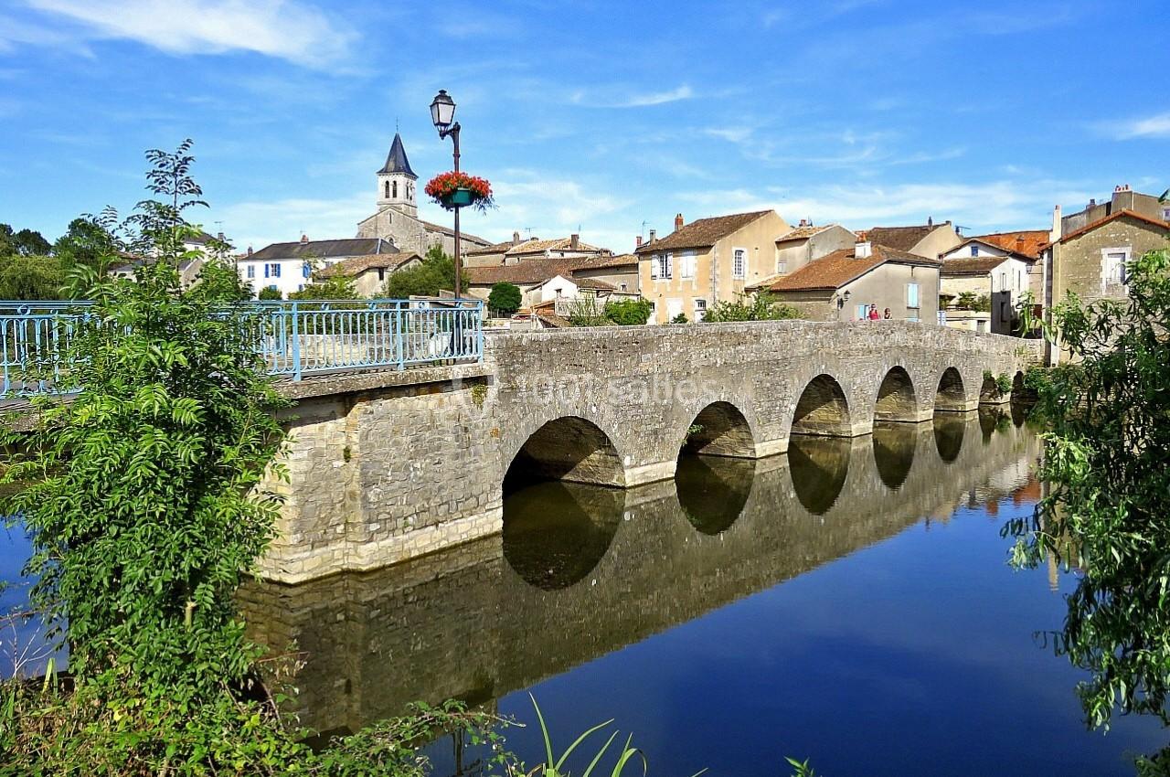 SANXAY Vue d'un pont en pierre à arches traversant une rivière, avec un village et un clocher en arrière-plan.