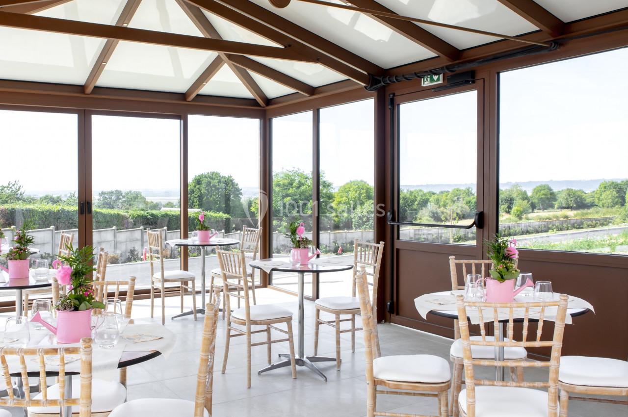 Location salle Saint-Samson-de-la-Roque (Eure) - Manoir Néo Normand #42 Salle lumineuse avec grandes baies vitrées, tables rondes décorées de fleurs roses et chaises en bois clair.