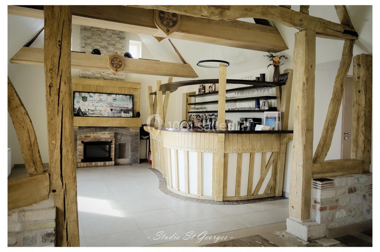 Location salle Saint-Samson-de-la-Roque (Eure) - Manoir Néo Normand #78 Comptoir en bois dans un espace lumineux avec poutres apparentes, cheminée et étagères garnies de bouteilles.