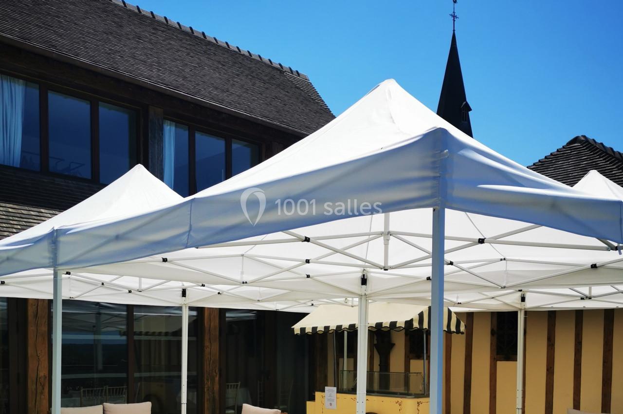 Location salle Saint-Samson-de-la-Roque (Eure) - Manoir Néo Normand #88 Tentes blanches installées sur une terrasse ensoleillée, entourées de mobilier d'extérieur et d'un bâtiment en arrière-plan.