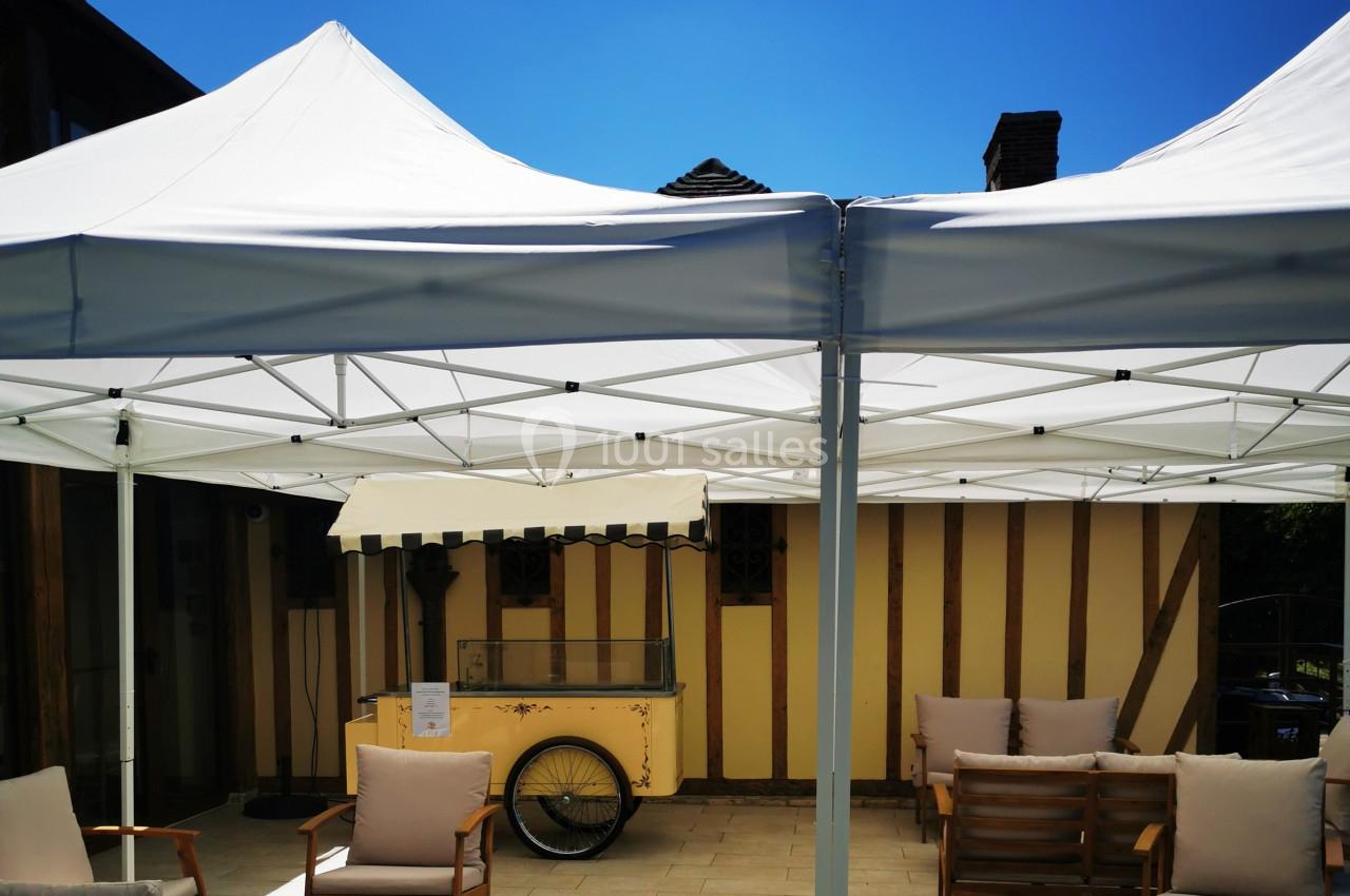 Location salle Saint-Samson-de-la-Roque (Eure) - Manoir Néo Normand #97 Tentes blanches installées sur une terrasse avec des fauteuils en bois et un chariot vintage en arrière-plan.