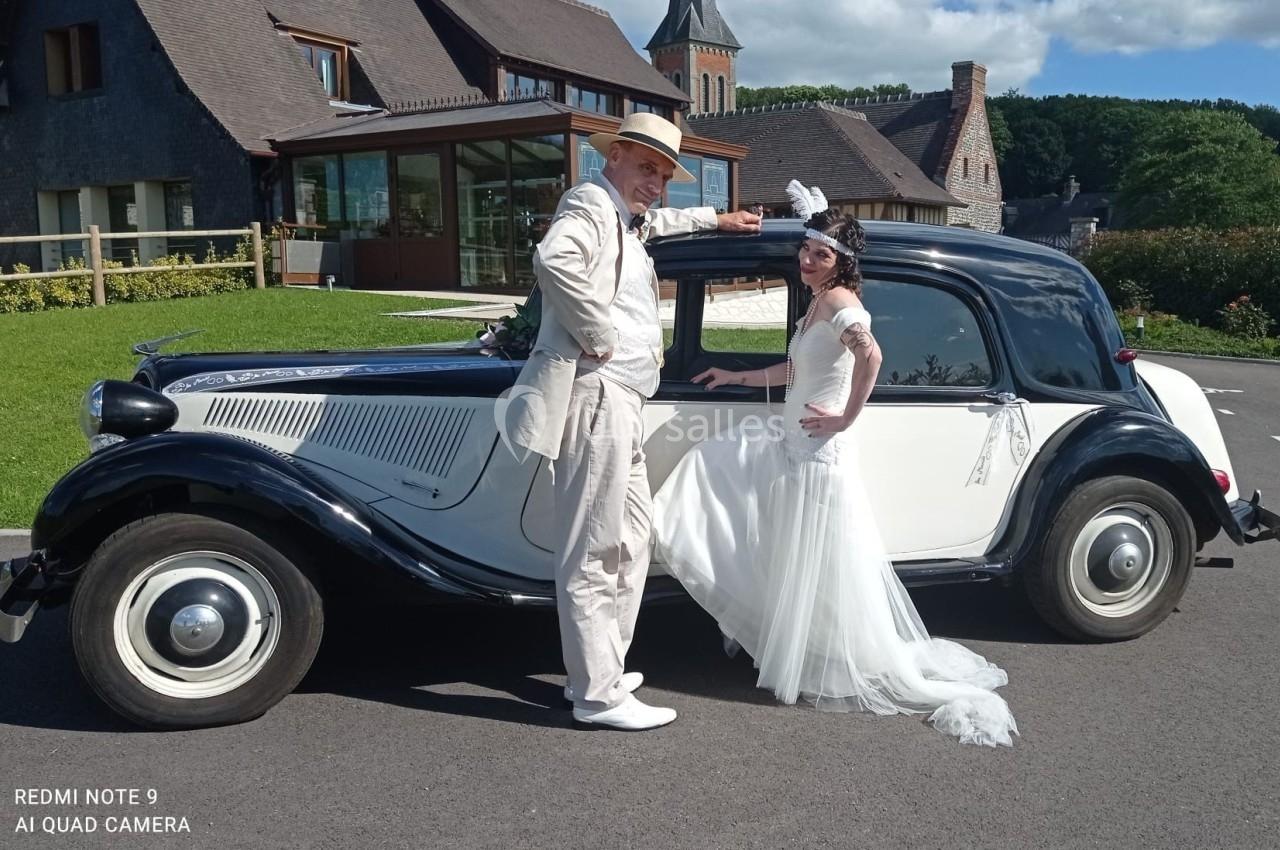 Location salle Saint-Samson-de-la-Roque (Eure) - Manoir Néo Normand #10 Un couple en tenue de mariage pose devant une voiture ancienne noire et blanche, sur une route près d'un bâtiment.