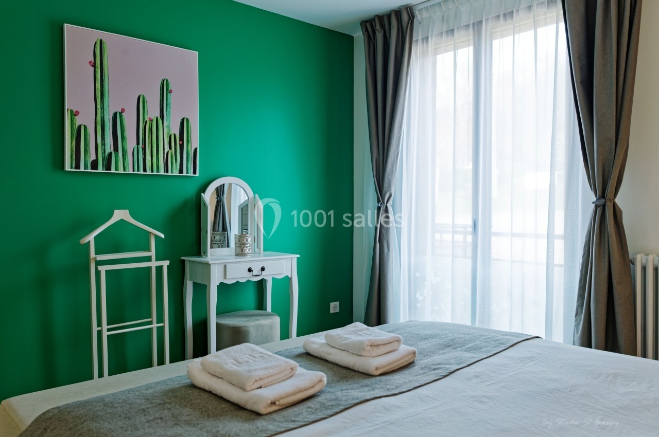 Chambre lumineuse avec mur vert, tableau de cactus, coiffeuse blanche, lit avec serviettes pliées et grande fenêtre avec…