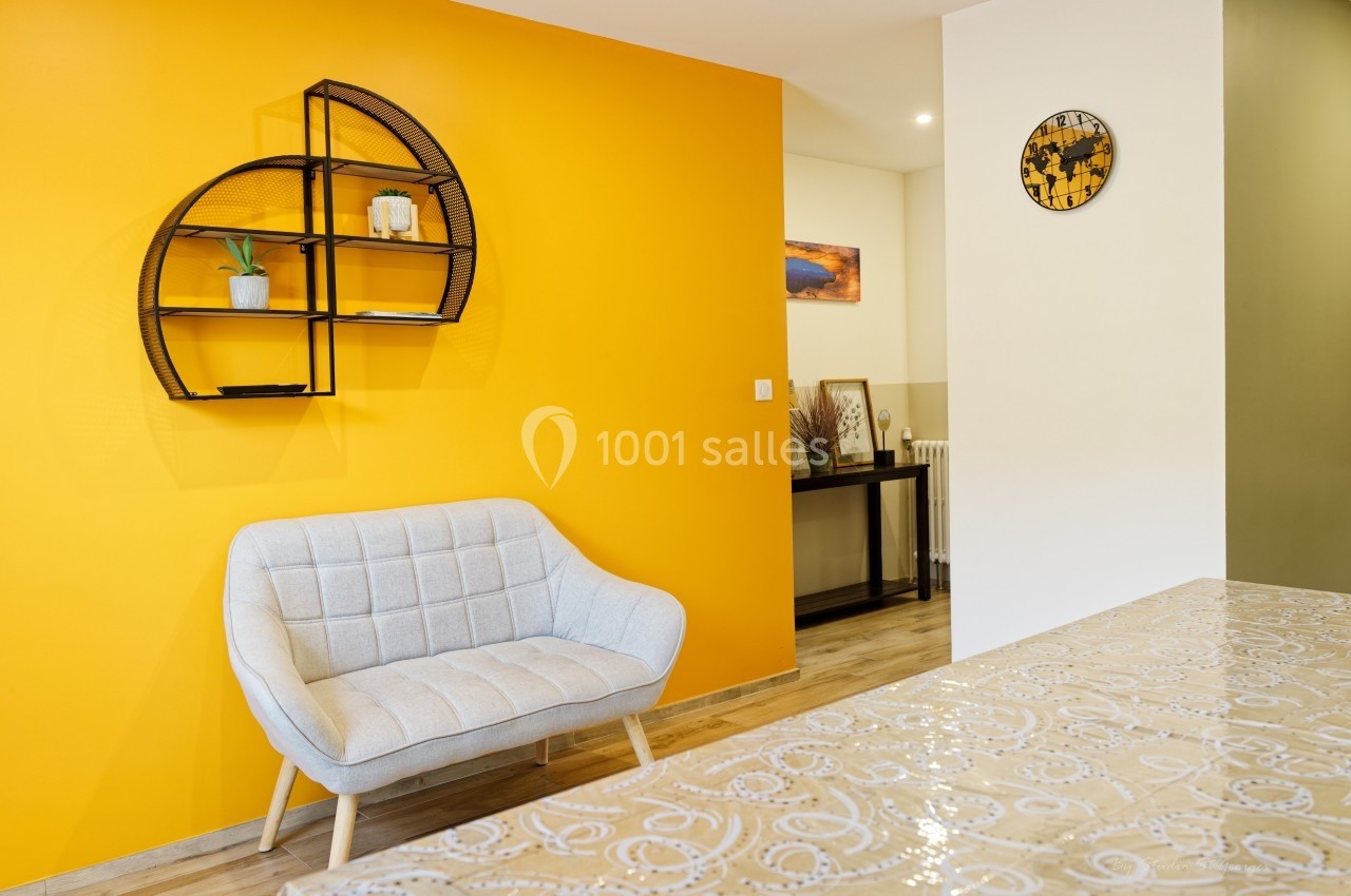 Mur jaune avec étagère décorative, fauteuil gris clair, table en premier plan et éléments de décoration en arrière-plan.