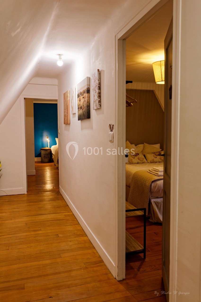 Couloir avec parquet menant à une chambre éclairée, décorée de tableaux et d'une lampe sur table.