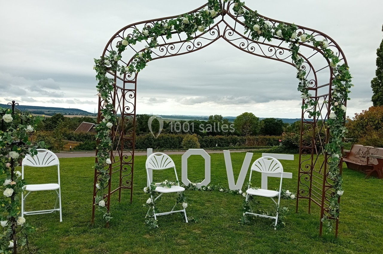 Arche décorée de feuillage et de fleurs blanches avec des chaises blanches sur une pelouse, vue sur la campagne.