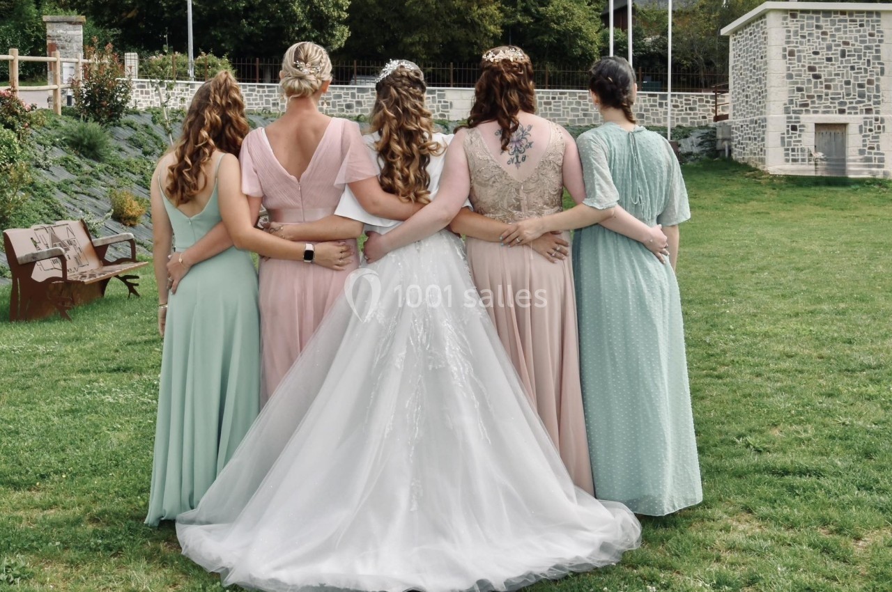 Cinq femmes de dos, dont une en robe de mariée, se tenant par la taille dans un jardin verdoyant.