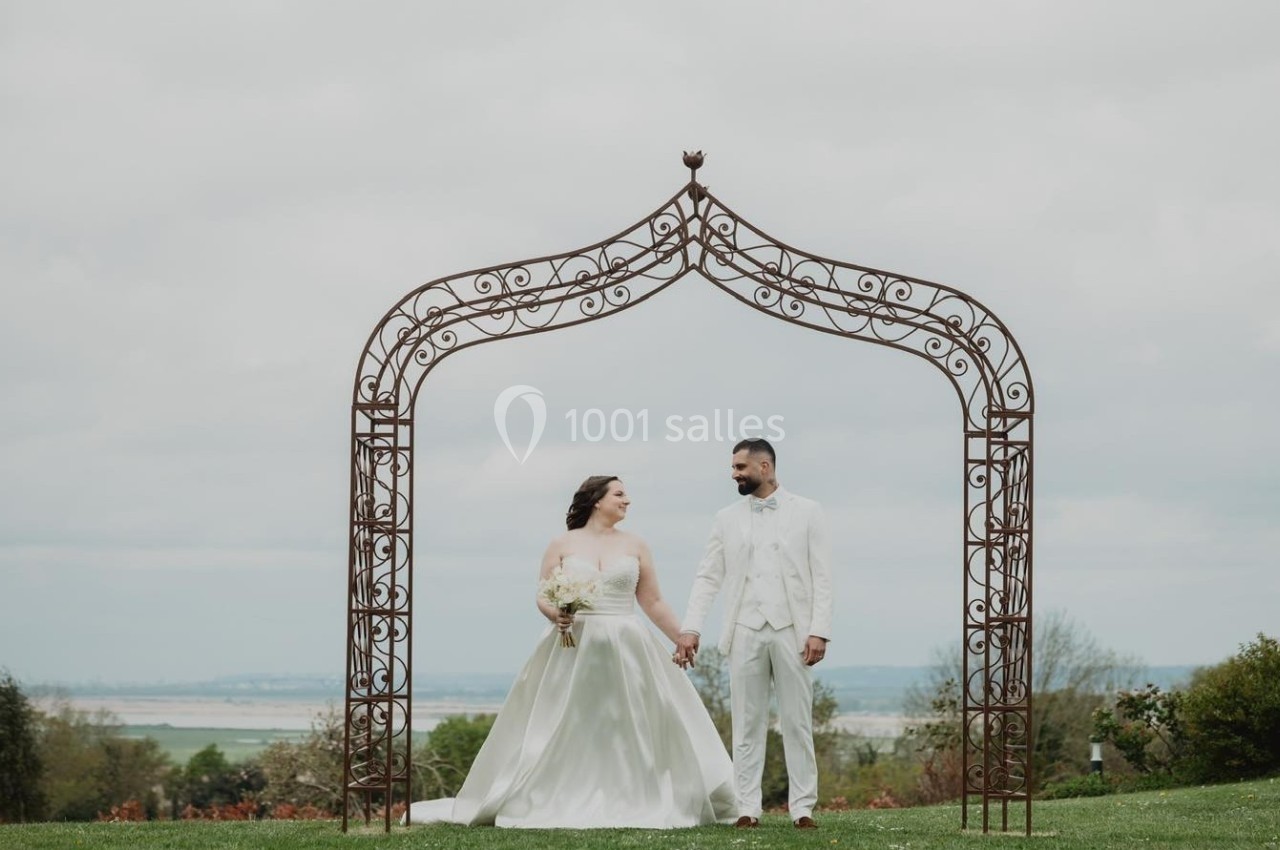 Un couple en tenue de mariage pose sous une arche métallique décorative dans un jardin avec vue dégagée.