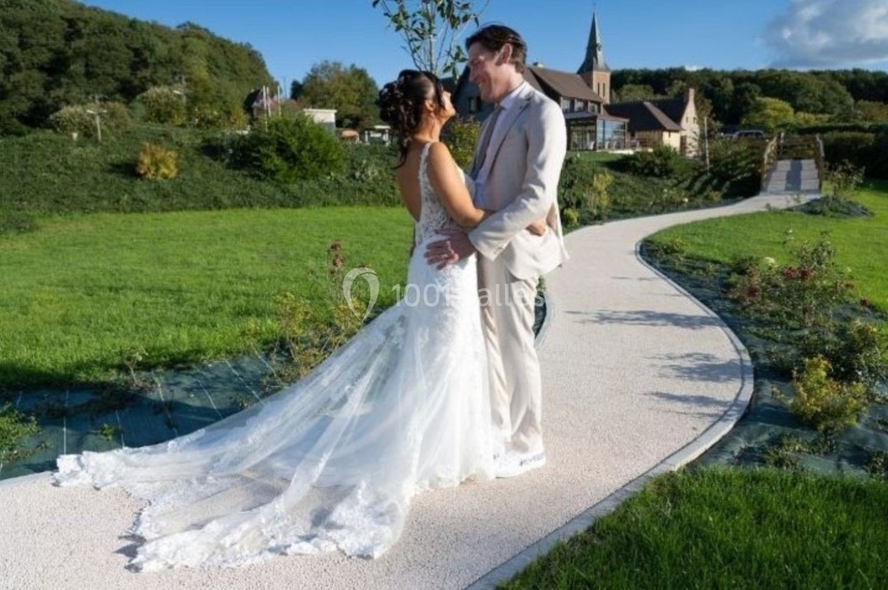 Un couple en tenue de mariage se tient sur un chemin bordé de verdure, avec une église en arrière-plan.
