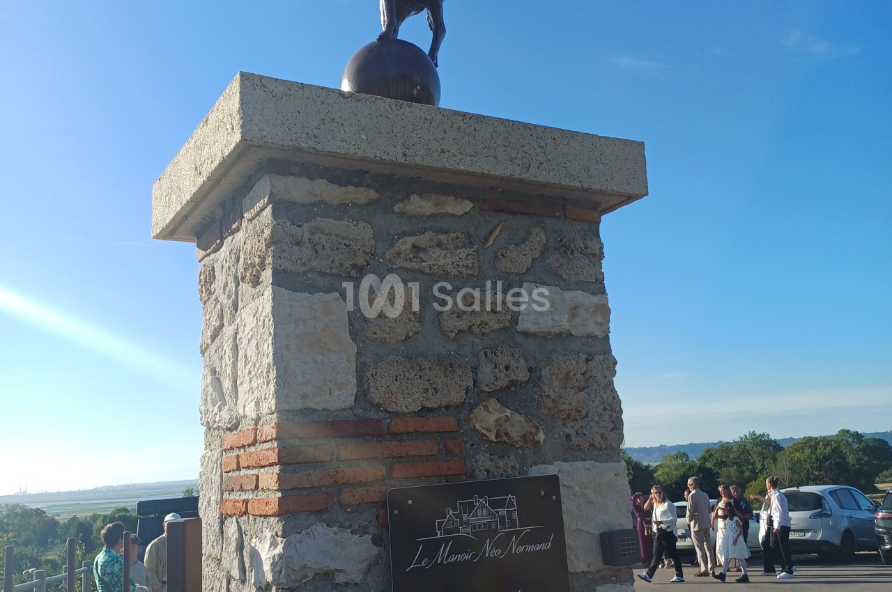 Colonne en pierre avec cloche métallique, vue sur vallée verdoyante et mer en arrière-plan.