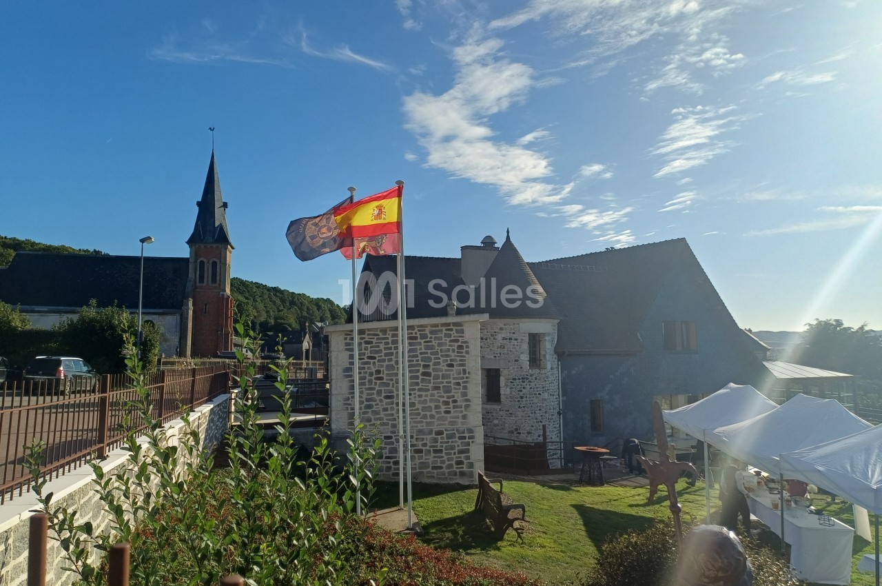 Manoir en pierre avec clocher d'église en arrière-plan, drapeau espagnol flottant et tente blanche visible à droite.