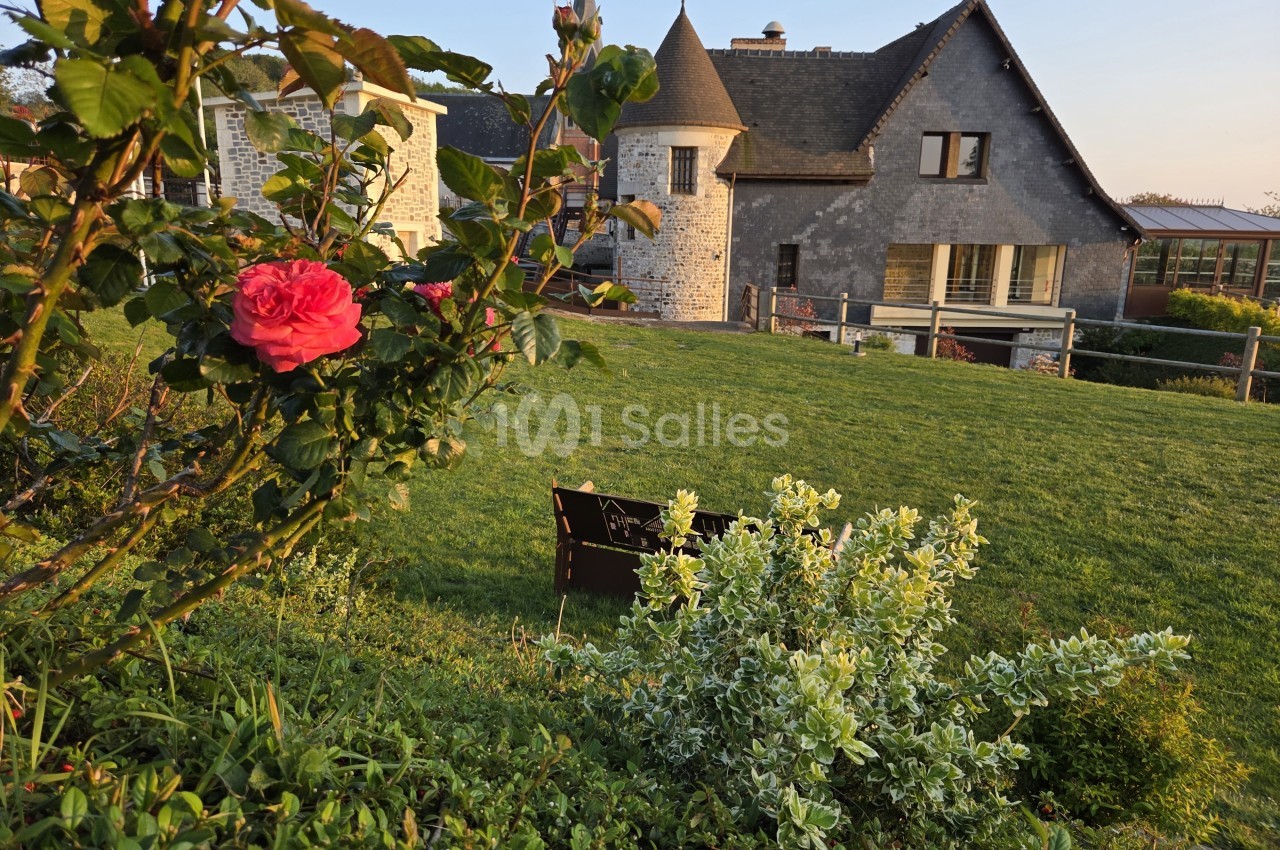 Manoir en pierre avec tourelle, parc arborisé avec roses et arbustes, chaises noires sur la pelouse.