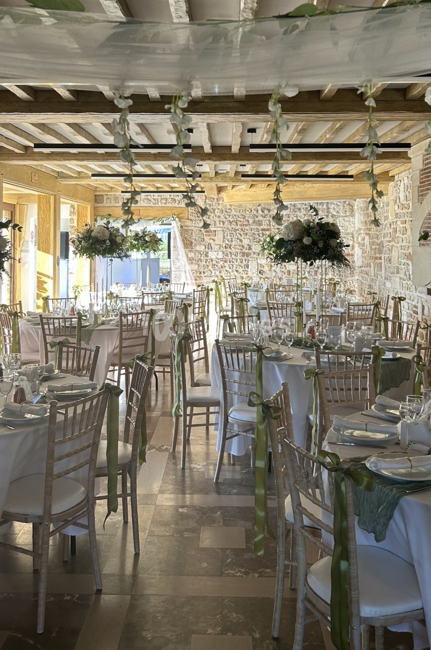 Salle de réception aux murs de pierre apparente et poutres en bois, dressée de tables rondes nappées de blanc, chaises en bois clair et rubans verts.