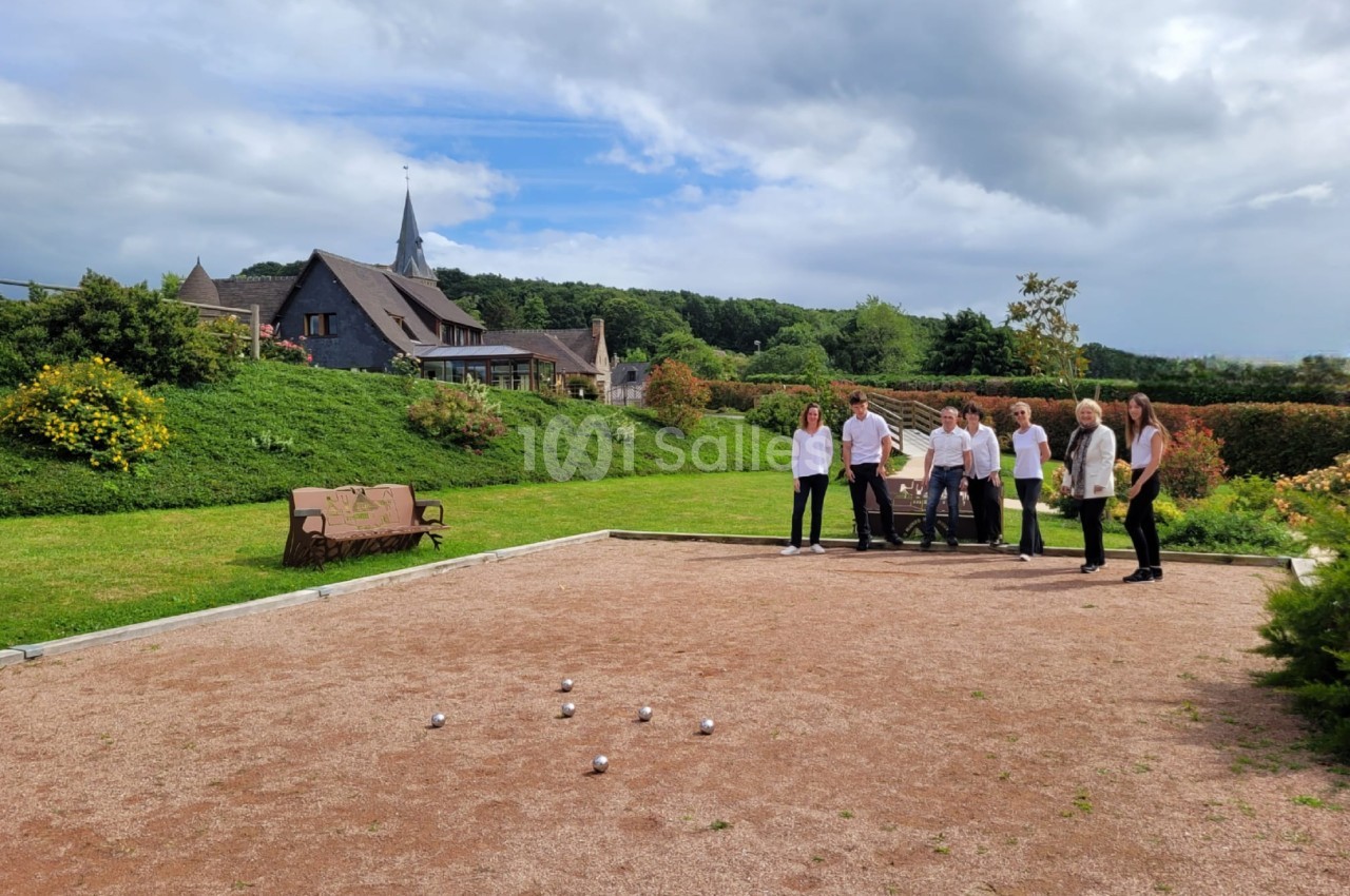 Terrain de pétanque en graviers au premier plan, avec six personnes en tenue blanche et noire debout près d'un banc en bois, devant une demeure à toit d'ardoise et un clocher. Let me check the constraints: one sentence, under 125 characters? That
