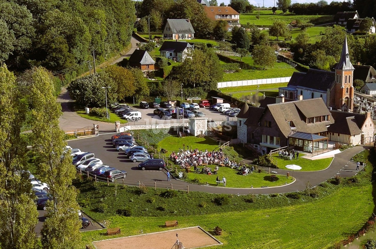Manoir néo-normand à colombages vu du ciel, avec des convives assis sur la pelouse en plein air et une église voisine.