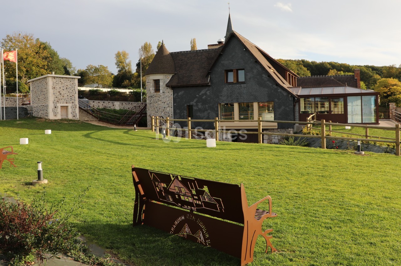 Façade en ardoise avec tourelle et véranda vitrée, vue depuis une pelouse et un banc en métal découpé au premier plan.