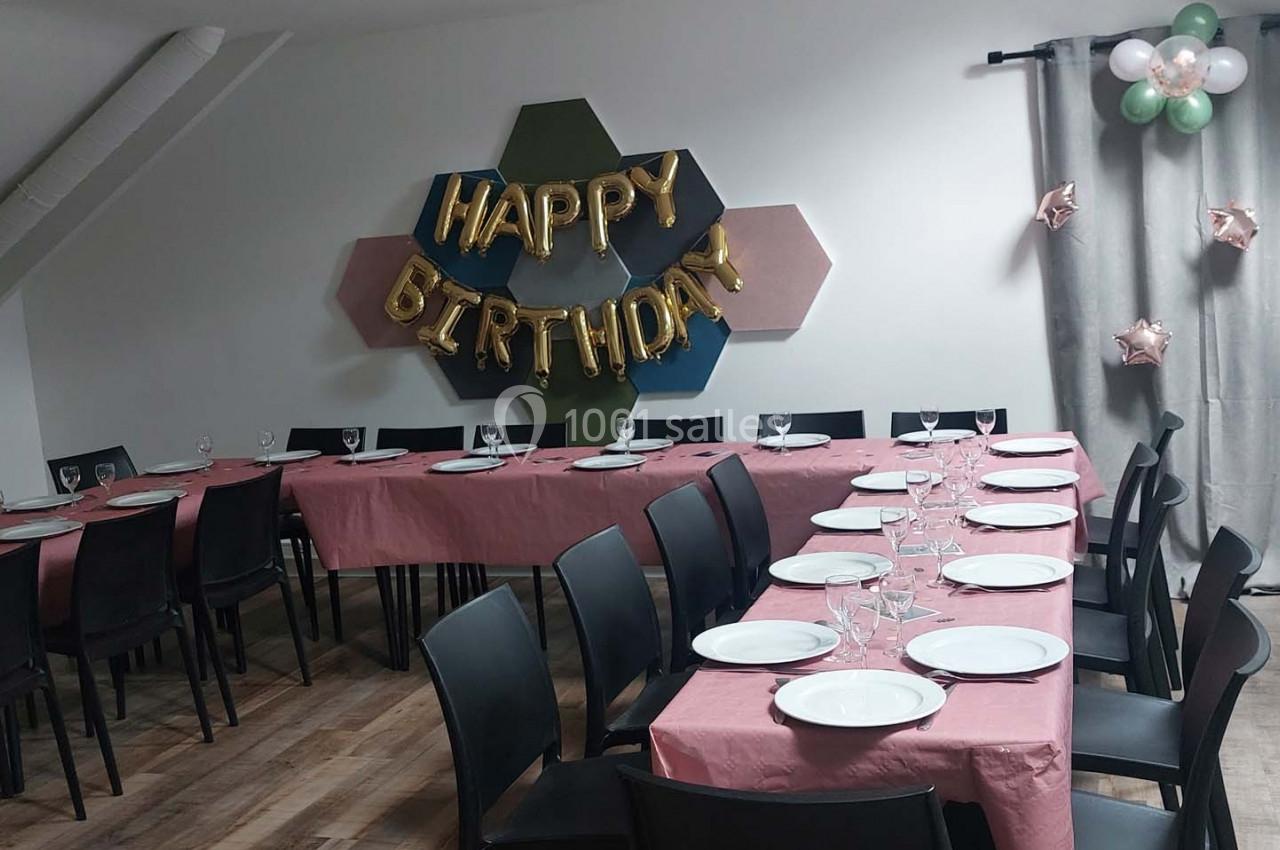 Salle décorée pour un anniversaire avec tables dressées, nappes roses, ballons et une bannière ’Happy Birthday’.