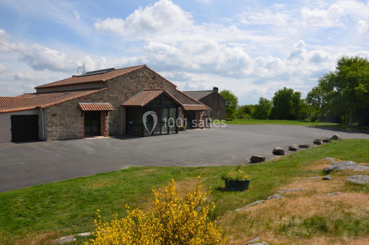 Bâtiment en pierre avec toit en tuiles, entouré d'une grande cour asphaltée et d'espaces verts parsemés de buissons.
