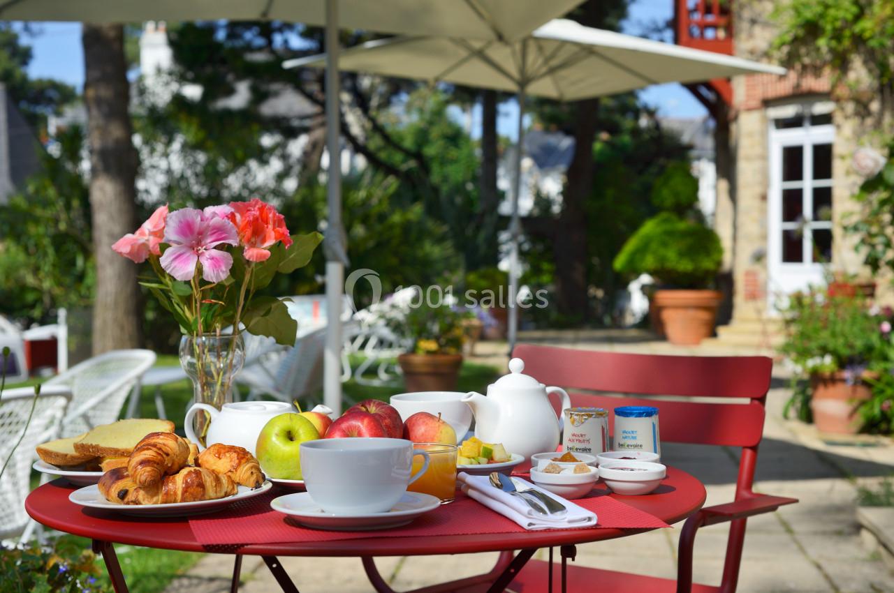 Petit-déjeuner servi en extérieur sur une table rouge avec viennoiseries, fruits, théière et fleurs en arrière-plan.