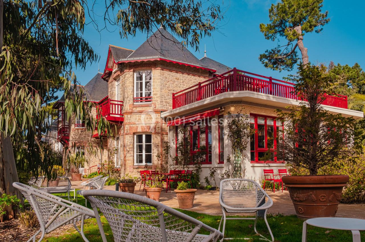 Maison en pierre avec balcons et volets rouges, entourée de végétation et de mobilier de jardin en métal blanc.