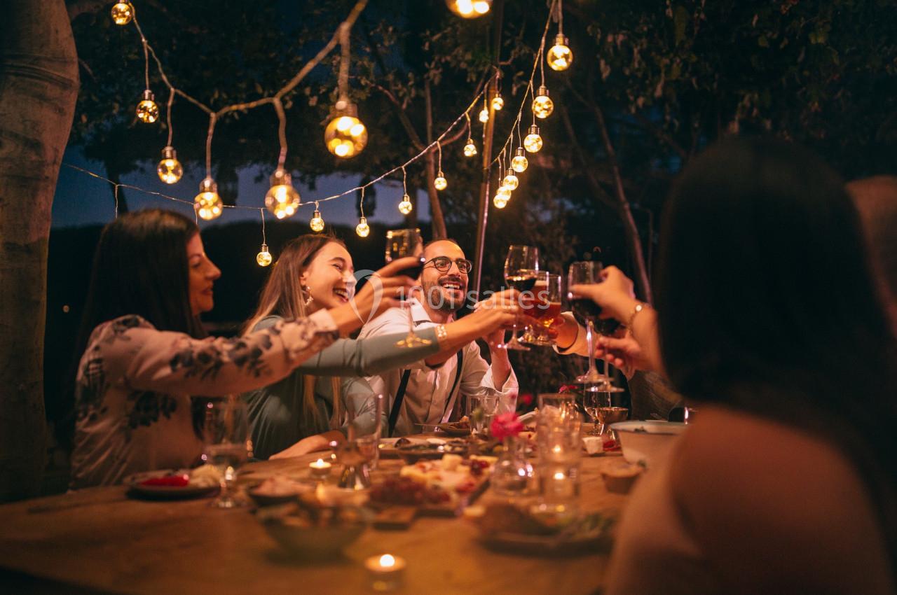 Un groupe de personnes trinque autour d'une table en extérieur, décorée de lumières suspendues, en soirée.