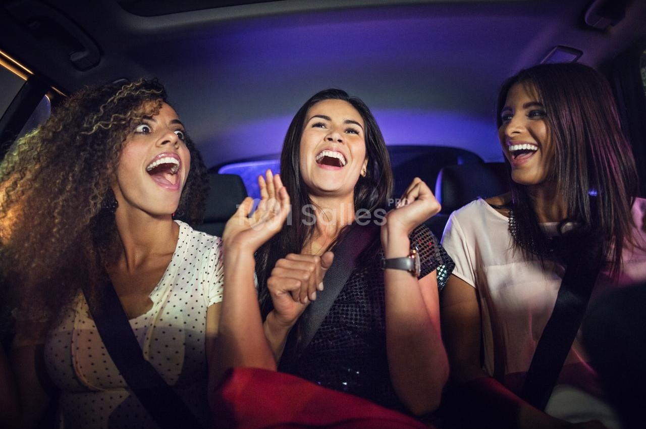 Trois femmes souriantes, assises dans une voiture, discutent et rient dans une ambiance nocturne.