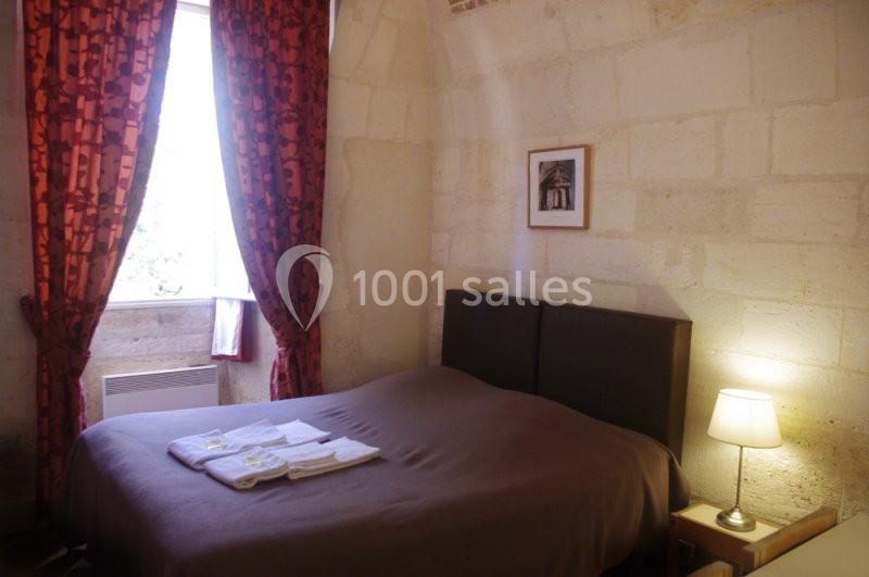 Chambre avec lit double, draps pliés, lampe allumée, mur en pierre, fenêtre avec rideaux rouges laissant passer la lumière.