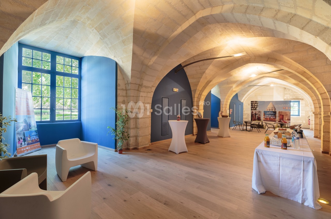Salle voûtée lumineuse avec murs en pierre, grandes fenêtres, mobilier moderne et table de collation dressée.