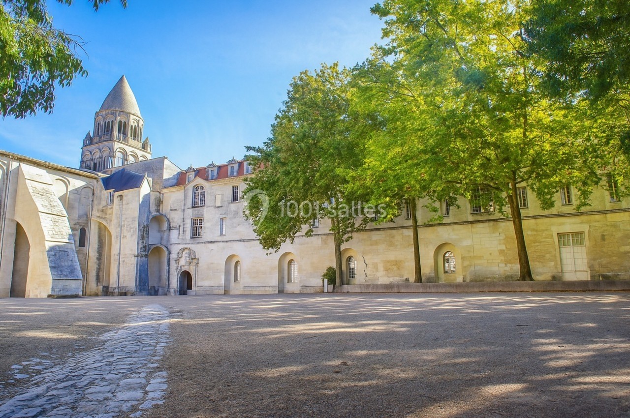 Cour pavée bordée d'arbres et de bâtiments historiques, avec un clocher en arrière-plan sous un ciel bleu.