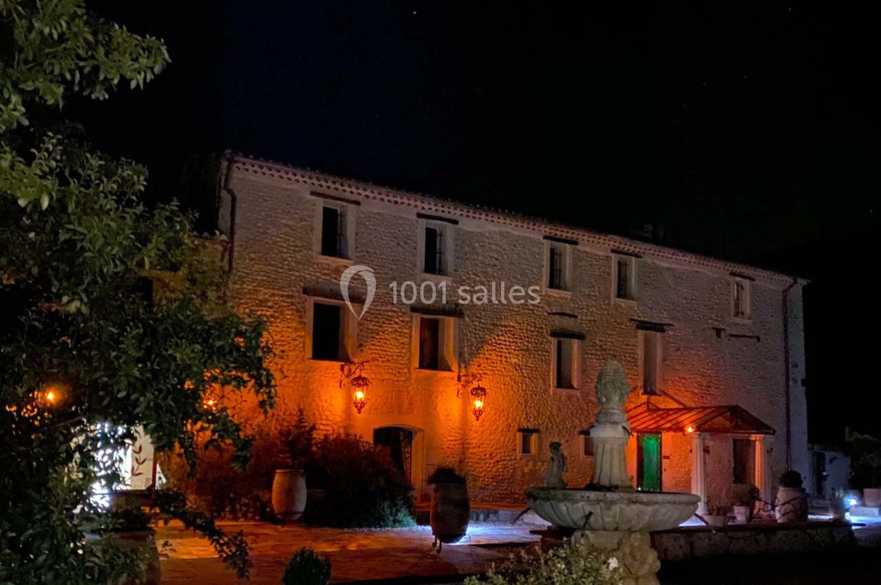Façade d'une bâtisse en pierre éclairée de nuit, avec une fontaine au premier plan et des lanternes allumées.