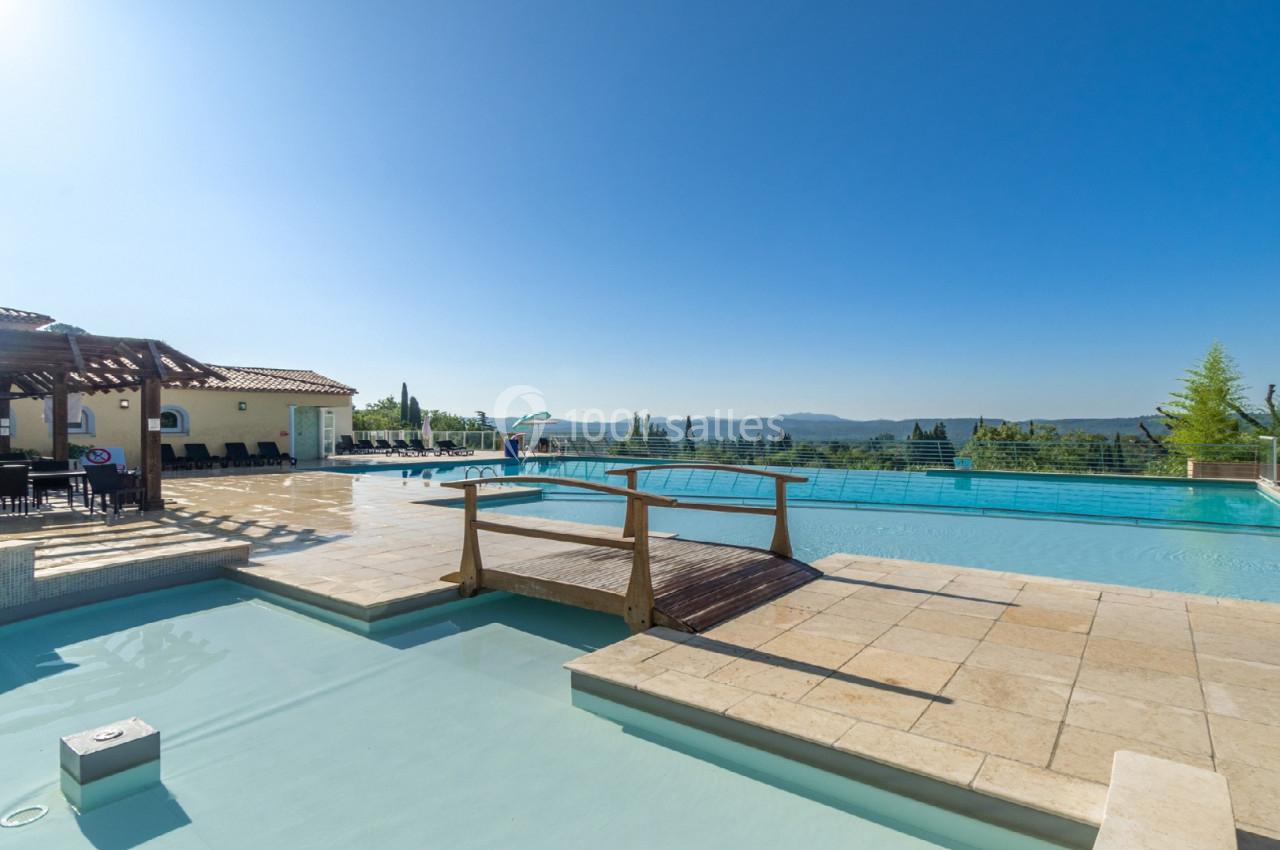 Piscine extérieure avec terrasse ensoleillée, pont en bois et vue panoramique sur un paysage vallonné.