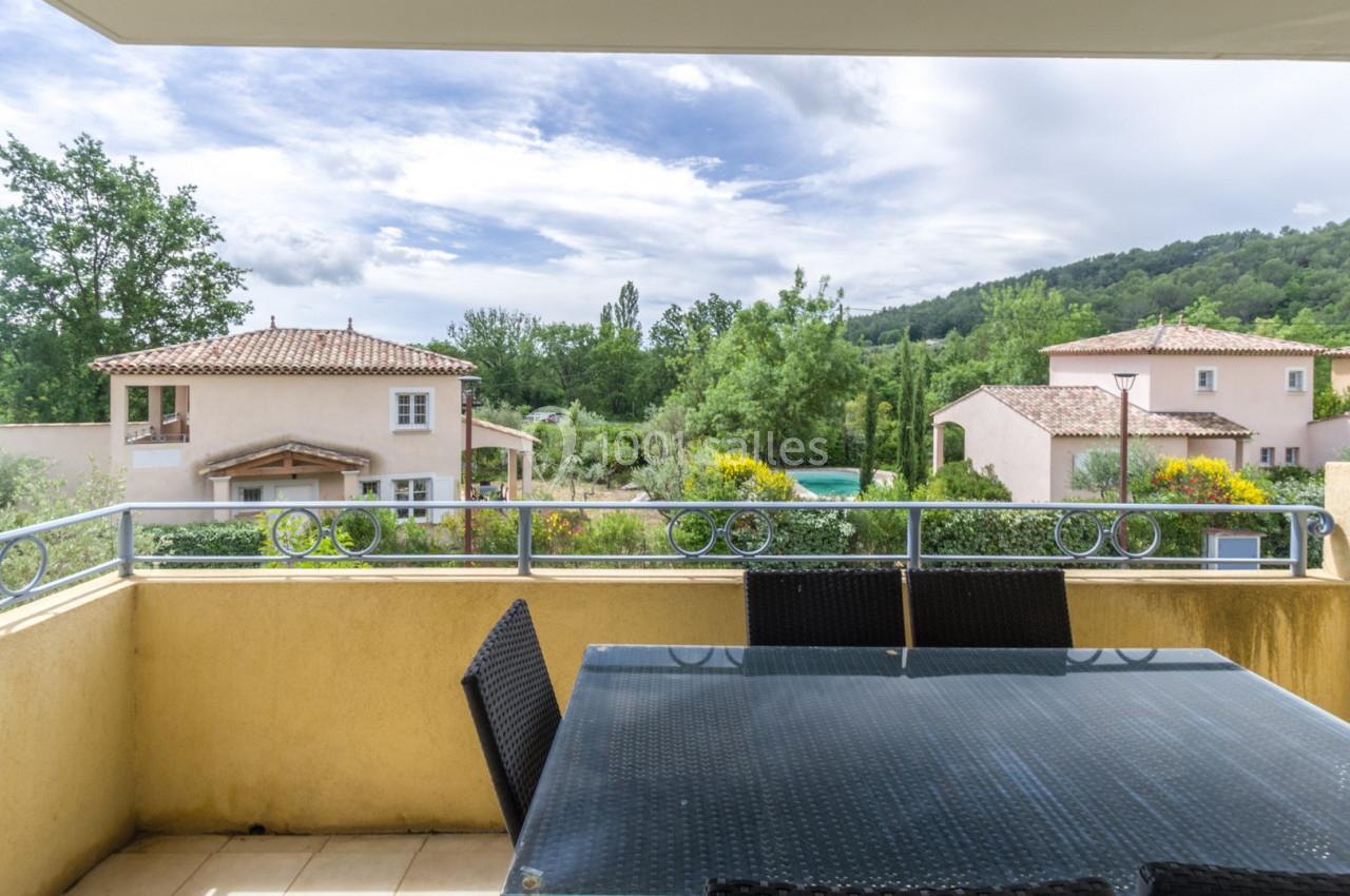 Vue depuis une terrasse avec table en verre et chaises, donnant sur des maisons, un jardin et des collines verdoyantes.