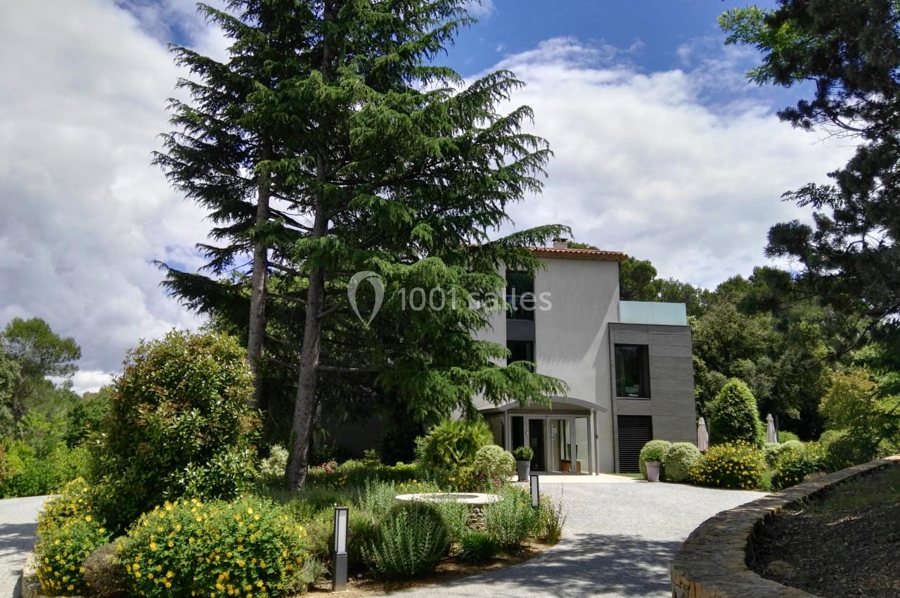Maison moderne entourée de végétation luxuriante, avec allée en gravier et arbres imposants sous un ciel partiellement…