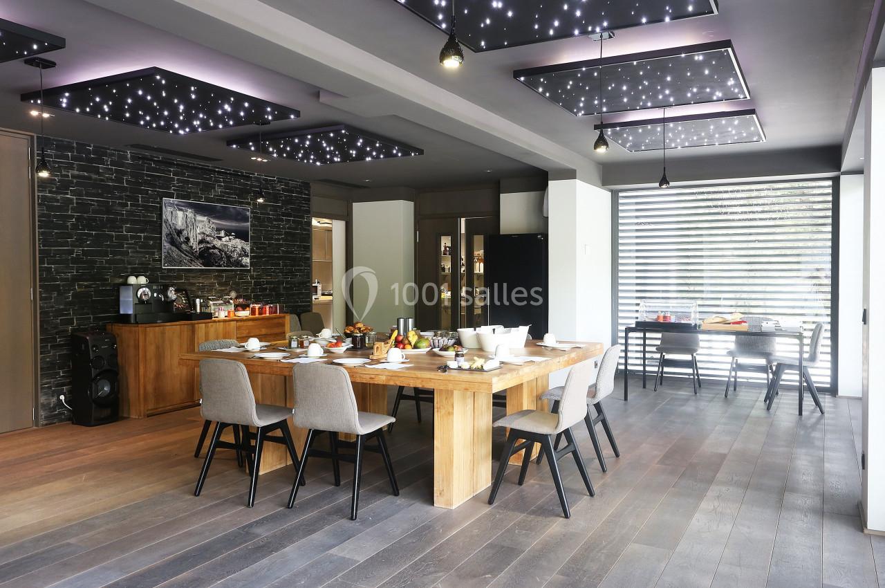 Salle à manger moderne avec grande table en bois, chaises grises, éclairage étoilé au plafond et cuisine ouverte en arrière…